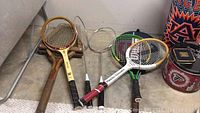 Five rackets arranged on the floor including two vintage wooden tennis rackets, a metal badminton racket, and two modern tennis rackets.