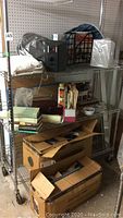 Metal shelving with various items including boxed JBL Control 24C speaker and unboxed JBL Control 26C ceiling speaker on top shelf alongside other miscellaneous items.