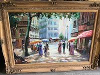 Frontal view of framed oil painting depicting a lively street scene with multiple figures, trees, and buildings.