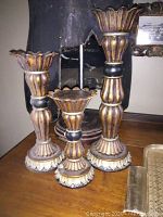 Front view of three carved ceramic candlesticks with floral scalloped edges on top and base, showcasing different heights and an antique dark brown and gold finish with black mid rings.