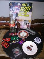 Photo showing 1982 NHL Hockey Sticker Album, Coca-Cola bottle, two plates, stapler, pin button and toy cars arranged on table.