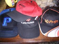 Group of four men's baseball caps showing logos of Kubota, Midas, Camar, and blue cap with 'P' logo laid out on wooden surface