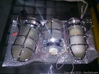 Three heavy industrial factory lights shown together on bubble wrap. Two are wall mount style with flat backplates, one is ceiling mount with round base. Chrome cage covers frosted glass cylinders.