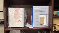 White Levoit air purifier unit next to its box on a shelf, sealed in plastic wrap, showing front and side.