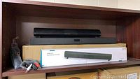 Two black soundbars displayed on a shelf including packaging box and cables.