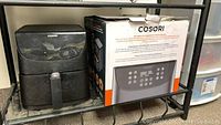Cosori air fryer with box on shelf, showing front and side of appliance and packaging box with control panel image.