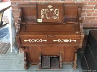 Front view of Eastlake style wooden pump organ showing decorative carved floral panel on music stand, inlaid front panel, and dual foot pedals.