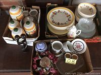 Photo showing assortment of dishes, canisters, ceramics, glassware, candle holders, and decorative dried flowers arranged in cardboard boxes on a table.