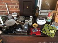 Photo showing an assortment of glassware, ceramics, electronic devices, decorative items, and a key holder arranged on a wooden surface with brick backdrop.