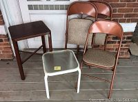 Photo showing 6 pieces: two brown metal folding chairs, two beige-patterned folding chairs, a small dark wooden side table, and a white metal garden table with glass top