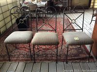 Three wrought iron chairs with brown upholstered seats placed side by side on a wooden floor against a paneled wall, showing their front and side profiles.