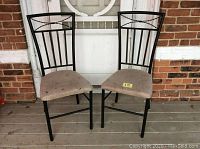 Pair of black metal frame chairs with beige upholstered seats placed on a porch against a brick wall and door.