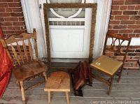 Photo showing Victorian picture frame, two wooden folding chairs, small wooden stool, and pine Arrow chair with beige cushion on a porch floor against a white door with brick wall background.