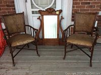 Two bamboo armchairs flank a wooden framed beveled mirror with carved details, set against a brick wall.