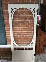 White wooden gingerbread screen door with decorative carved wooden trim and spindles. Door is upright leaning against a brick wall.
