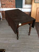Photo of a vintage dark stained pine drop leaf farm table with turned legs and two drop leaves folded down, placed on a wooden deck.