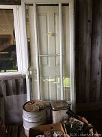 Photo of the white framed full glass storm door leaning against wall with two large metal oil bins and two boxes of various items on floor.
