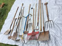 All garden tools laid out on a white tarp on grass showing full length and heads, including rakes, forks, and shovels.