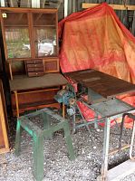 Overview of vintage kitchen cabinet with glass doors and fold-up table next to vintage saw table and metal base with missing blade.