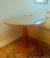 Round wooden side table base with thick glass top resting on it, placed against a wall.