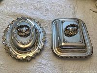 Two silver-plated vegetable dishes with ornate lids, one oval and one rectangular, showing decorative edges and handles