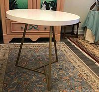 Side angle showing off-white glossy round tabletop and brass geometric metal base on patterned rug