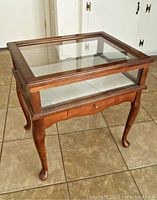 Display case with wooden frame and glass lid, showing front view with drawer and curved legs.