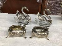 Overview of both swan dishes with spoons and two floral-embossed footed pots with spoons displayed on towel.