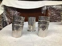 Seven silver shot glasses displayed on a white cloth. Six have detailed leaf or feather engravings, one plain with the text "SHOT" on it.