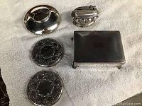 Top-down view of two round compact mirrors, silver-tone inkwell, lighter, and square trinket box on white towel