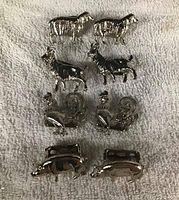 Four pairs of silver-tone animal place card holders arranged on towel surface. Pairs include sheep, goats, roosters, and pigs. Each figure has card holding slots on the back.
