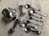 Overview of 4 baby spoons, 3 baby forks, 2 rattles and baby cup on a neutral fabric background.