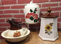 Photo showing McCoy ceramic items including a bowl and pitcher set with pomegranate designs, a cylindrical cookie jar with red poppies design, and a hexagonal cookie jar with sunflower design and brown lid.