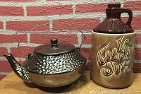 Two cookie jars, one shaped like a tea kettle with hammered metal finish and the other a brown jug with embossed lettering.