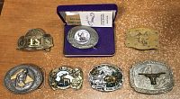 Photo showing all seven belt buckles arranged on a table, one is boxed and new, others show various wildlife and western themed designs.