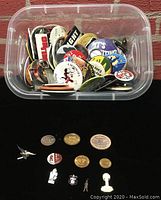 Top-down view showing a clear plastic bin filled with assorted round and shaped pins, keychains, and a variety of tokens arranged on a flat black surface below.