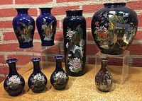Lot of nine blue vases with various hand-painted traditional Japanese designs, against a brick wall background.