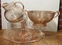 Set of three pink Depression glass pieces: ball pitcher, fruit bowl, tidbit plate, all shown together on a surface