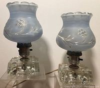 Two vintage boudoir lamps with clear glass bases and blue frosted pressed glass floral shades shown from front view.