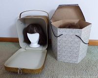Photo showing gold travel wig box open with white Styrofoam head and brown wig inside, and tall gray-patterned box with beige Styrofoam head and wig inside