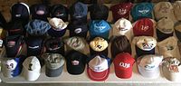 Photo showing 33 baseball caps arranged on table in multiple colors including blue, red, beige, black, white, and teal with various logos.
