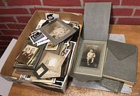 Box full of old photographs, some framed portraits visible on top and a photo holder folder next to box
