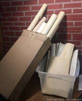 Rolled and loose blueprints in cardboard box and plastic bin leaning against brick wall.