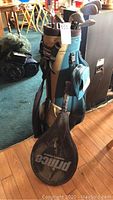 Golf bag standing upright on hardwood floor with tennis racket cover hanging on side, showing overall condition and color of bag.