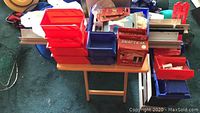 View of red and blue small organizing bins, shelving brackets, boxed hardware, and assorted items on tables and floor area.