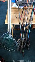 Multiple fishing rods standing together with two fishing nets leaning on them. Rods vary in length and color, mesh nets one larger, one smaller with metal handles.