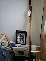 Two wooden hockey sticks standing upright against a wall beside a framed signed hockey photograph on floor and a white seat cushion with a bulldog graphic.