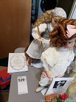 Photo of two porcelain dolls with papers and stands, one dressed as Cinderella, the other as The Little Girl with a Curl.