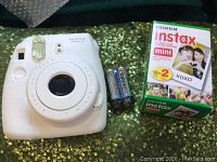 White Fujifilm Instax Mini 8 instant camera with two new AA batteries and pack of Fujifilm Instax Mini instant film on green sequin fabric.