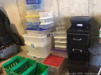 Cluster of assorted bins leaning against concrete wall, showing clear plastic bins at left, green crates front, red-lidded bin and black metal bin stack at right
